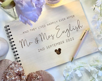 Puede incluir: Un libro de visitas de madera con el texto "...and they lived happily ever after Mr & Mrs English 2nd September 2022" y una silueta de Mickey Mouse. El libro está abierto y tiene una encuadernación en espiral. Un bolígrafo rosa está sobre el libro.