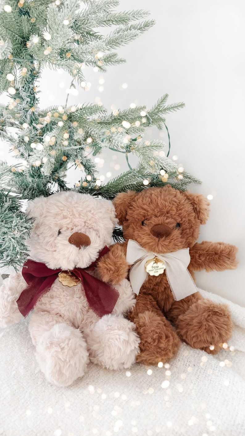 May include: Two plush teddy bears, one cream and one brown, each wearing a ribbon around its neck. The cream bear has a dark red ribbon and a gold-toned tag, while the brown bear has a white ribbon and a gold-toned tag with the text Ozzy Merry Christmas 2024.