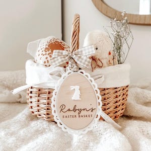 May include: A white wicker basket with a white and grey gingham ribbon and a wooden tag that says "Robyn's Easter Basket" with a bunny silhouette. The basket is filled with a stuffed animal, a brown and white patterned egg, and white flowers.