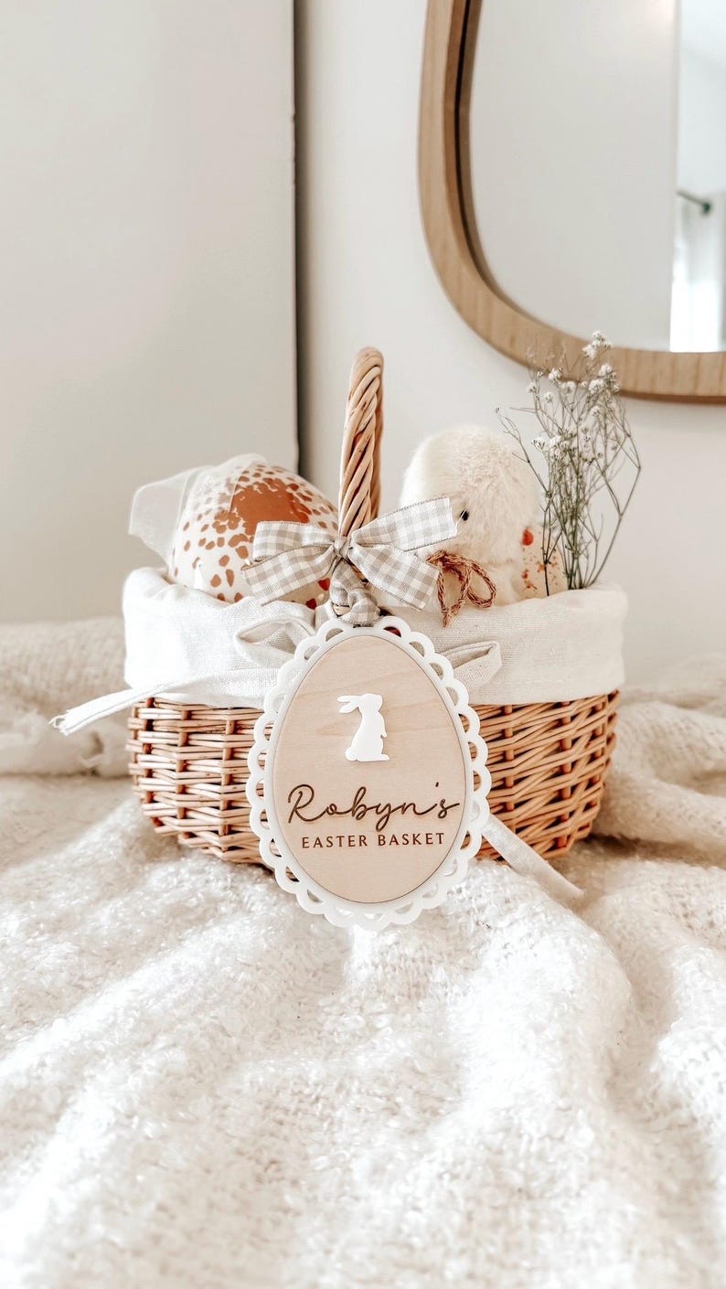 May include: A white wicker basket with a white and grey gingham ribbon and a wooden tag that says "Robyn's Easter Basket" with a bunny silhouette. The basket is filled with a stuffed animal, a brown and white patterned egg, and white flowers.