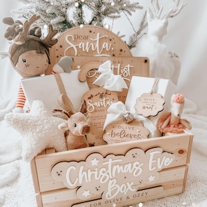 May include: A wooden crate with the words "Christmas Eve Box" and "For Olive & Ozzy" on it. The crate is filled with various items, including a stuffed reindeer, a wooden sign that says "Dear Santa", and a wooden ornament that says "Olive believes".