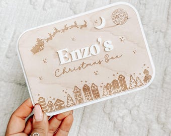 Puede incluir: Caja de Navidad de madera con borde blanco, con el nombre "Enzo's" en letras blancas en relieve. La caja está decorada con un trineo de Papá Noel, una luna, estrellas y una escena de pueblo. Las palabras "Christmas Box" están escritas en cursiva.