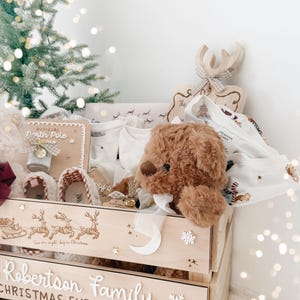 May include: A wooden Christmas Eve box for the Robertson family, featuring engraved reindeer, a village scene, and the text 'Robertson Family CHRISTMAS EVE BOX'. The crate contains a brown teddy bear, white clothing, and a North Pole sign.