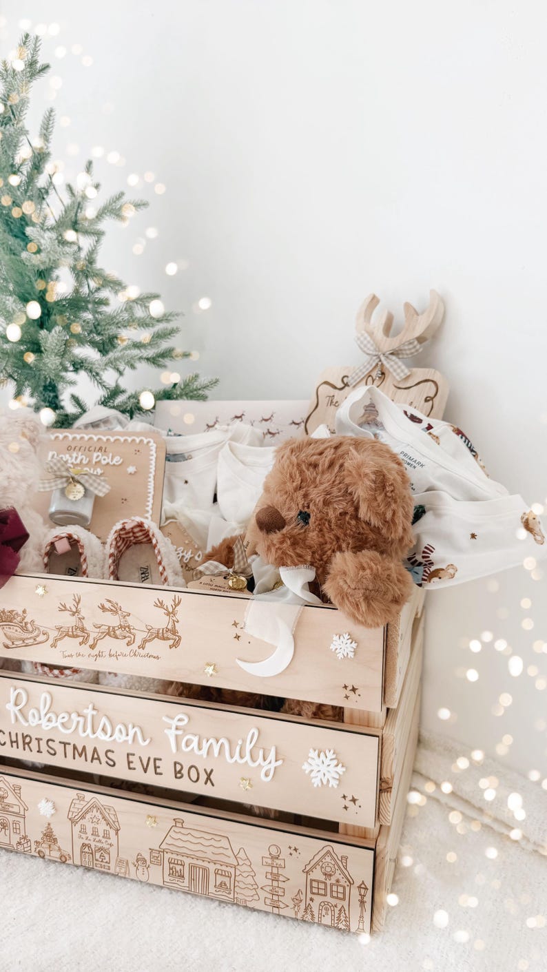 May include: A wooden Christmas Eve box for the Robertson family, featuring engraved reindeer, a village scene, and the text 'Robertson Family CHRISTMAS EVE BOX'. The crate contains a brown teddy bear, white clothing, and a North Pole sign.