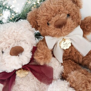May include: Two plush teddy bears, one cream and one brown, each wearing a ribbon and a gold-toned scalloped metal tag. The cream bear's tag reads Amelie's First Christmas 2024, and the brown bear's tag reads Ozzy Merry Christmas 2024.