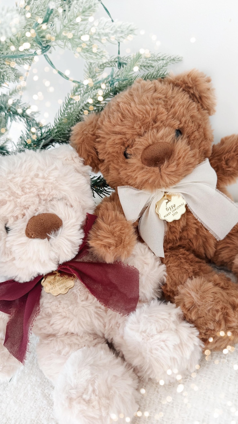 May include: Two plush teddy bears, one cream and one brown, each wearing a ribbon and a gold-toned scalloped metal tag. The cream bear's tag reads Amelie's First Christmas 2024, and the brown bear's tag reads Ozzy Merry Christmas 2024.