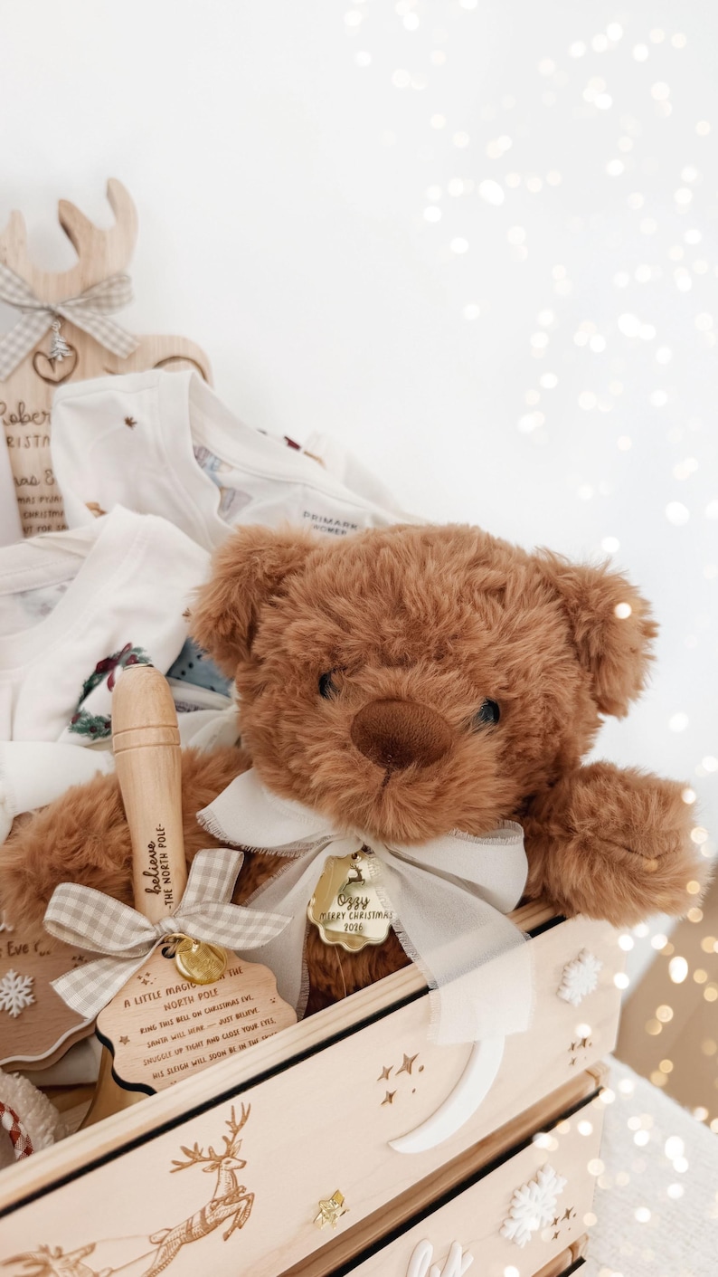 May include: A brown plush teddy bear with a white ribbon and a gold tag reading Ozzy Merry Christmas 2026, placed in a wooden crate with a bell featuring the text Believe The North Pole and A Little Magic From The North Pole.