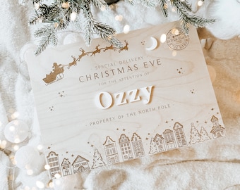 May include: A wooden sign with the text "SPECIAL DELIVERY CHRISTMAS EVE FOR THE ATTENTION OF Ozzy PROPERTY OF THE NORTH POLE" and a depiction of Santa Claus in a sleigh with reindeer flying over a town with houses and a Christmas tree.