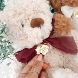 May include: A cream-colored plush teddy bear with a dark brown nose and a deep red ribbon tied around its neck. A gold-toned scalloped metal tag hangs from the ribbon, engraved with a reindeer icon and the text Amelia's FIRST CHRISTMAS 2026.