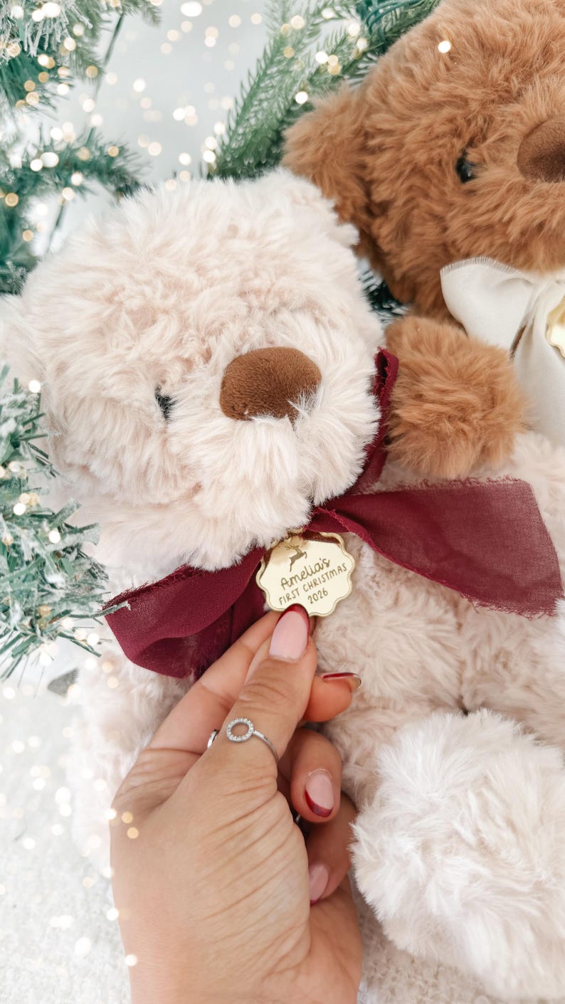 May include: A cream-colored plush teddy bear with a dark brown nose and a deep red ribbon tied around its neck. A gold-toned scalloped metal tag hangs from the ribbon, engraved with a reindeer icon and the text Amelia's FIRST CHRISTMAS 2026.