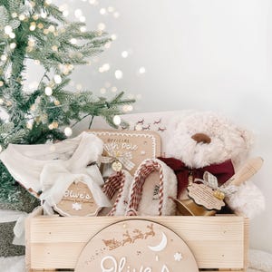 May include: A wooden crate labeled Olive's Christmas Eve Box with a graphic of a sleigh and village. Inside are white pajamas, a beige teddy bear with a maroon bow, plaid slippers, and a wooden tag reading Official North Pole.