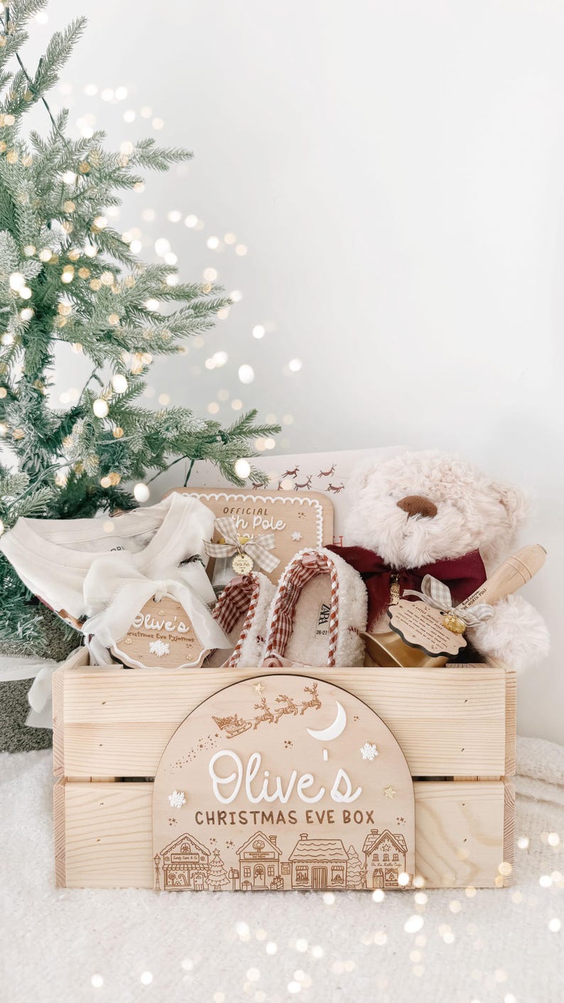 May include: A wooden crate labeled Olive's Christmas Eve Box with a graphic of a sleigh and village. Inside are white pajamas, a beige teddy bear with a maroon bow, plaid slippers, and a wooden tag reading Official North Pole.