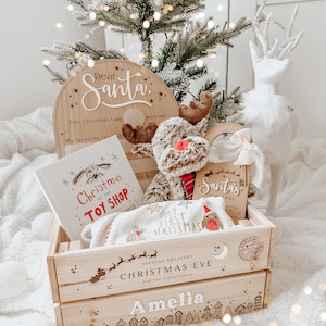May include: A wooden crate filled with Christmas-themed items. Includes a "Dear Santa" sign, a plush reindeer, a book titled "Christmas at the Toy Shop", and a blanket. The crate is personalized with the name "Amelia" and "Christmas Eve" text.