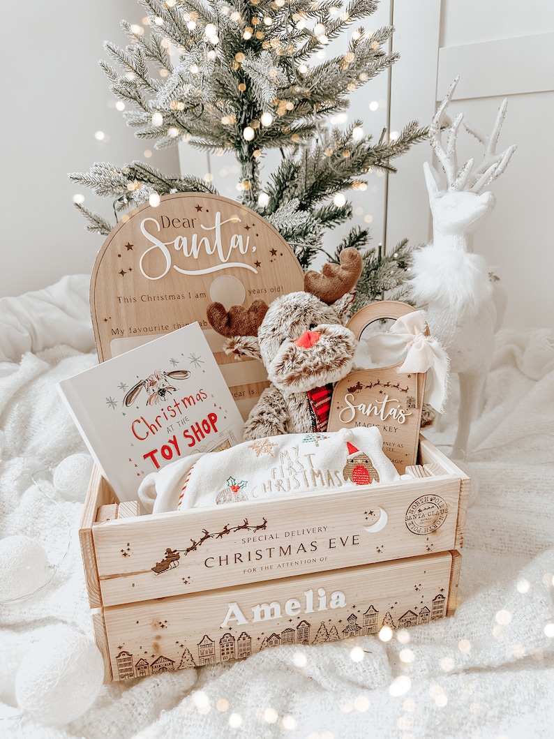May include: A wooden crate filled with Christmas-themed items. Includes a "Dear Santa" sign, a plush reindeer, a book titled "Christmas at the Toy Shop", and a blanket. The crate is personalized with the name "Amelia" and "Christmas Eve" text.