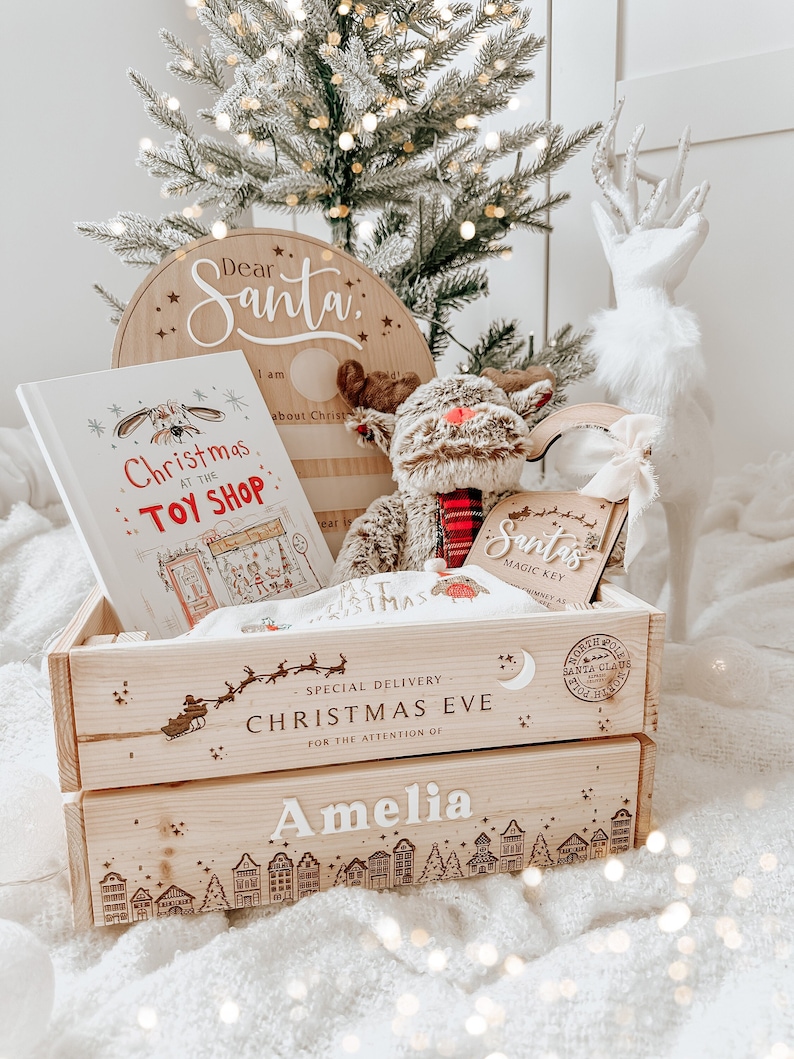 May include: A wooden Christmas Eve gift crate with the name "Amelia" on it, filled with a plush reindeer, a book titled "Christmas at the Toy Shop", and a "Dear Santa" sign. The crate is decorated with a town silhouette.