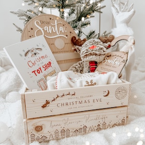 May include: A wooden Christmas Eve gift crate with a reindeer plush, a children's book titled "Christmas at the Toy Shop", and a "Dear Santa" sign. The crate is personalized with a name and a townscape design.