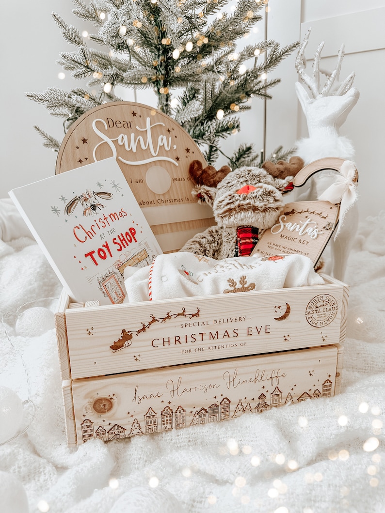 May include: A wooden Christmas Eve gift crate with a reindeer plush, a children's book titled "Christmas at the Toy Shop", and a "Dear Santa" sign. The crate is personalized with a name and a townscape design.
