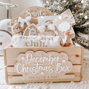 May include: A wooden Christmas box with the text "December Christmas Box" and "For Olive & Ozzy". The box contains a variety of items, including a plush bear, a rolled-up blanket with "Nice List Club" embroidered on it, and a wooden sign that says "Dear Santa".