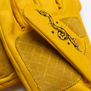 May include: Close-up of a pair of yellow leather gloves. The gloves have a quilted design on the palm and a black snake illustration with stars on the back of the hand. The gloves are made of a soft, supple leather.