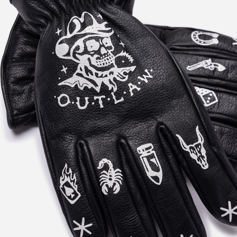 May include: Black leather gloves with white graphic designs. The gloves feature a skull wearing a cowboy hat, the word "OUTLAW", and other western-themed illustrations like a horseshoe, gun, and dice.