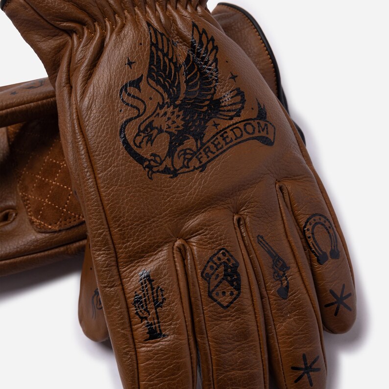 May include: Brown leather gloves with black tattoo-style graphics. The designs include an eagle with the word "FREEDOM", a cactus, dice, a revolver, a horseshoe, and stars. The gloves have a classic biker style.