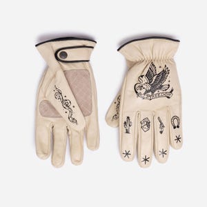 May include: A pair of cream-colored leather gloves with black trim and decorative tattoos. The right glove features an eagle with the word "FREEDOM". Other tattoos include a snake, cactus, dice, gun, horseshoe, and stars.