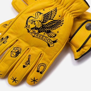 May include: Yellow leather gloves with black illustrations. The gloves feature an eagle with the word "FREEDOM", dice, a gun, a horseshoe, and stars. The gloves have black trim and a button closure.