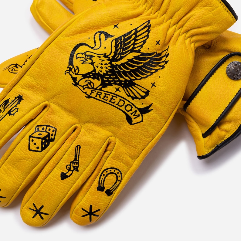 May include: Yellow leather gloves with black illustrations. The gloves feature an eagle with the word "FREEDOM", dice, a gun, a horseshoe, and stars. The gloves have black trim and a button closure.