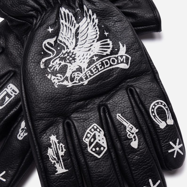 May include: Black leather gloves with white graphic designs. The gloves feature an eagle with the word "FREEDOM", a cactus, dice, a revolver, and a horseshoe. The gloves are designed for protection and style.