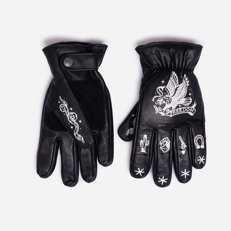 May include: Black leather gloves with white graphic designs. One glove features a snake design, while the other has an eagle with the word "FREEDOM" and other symbols. The gloves have a button closure.