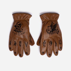 May include: A pair of brown leather gloves with black tattoo-style designs. One glove features a skull wearing a cowboy hat with the word "OUTLAW". The other glove has an eagle with the word "FREEDOM". Additional small tattoos are on the fingers.