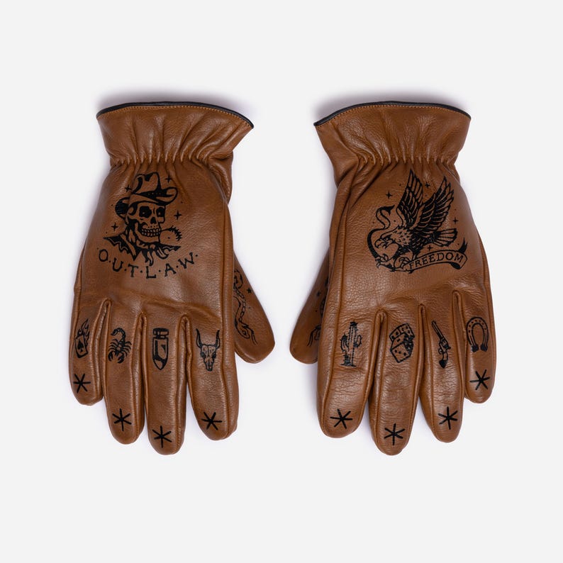 May include: A pair of brown leather gloves with black tattoo-style designs. One glove features a skull wearing a cowboy hat with the word "OUTLAW". The other glove has an eagle with the word "FREEDOM". Additional small tattoos are on the fingers.