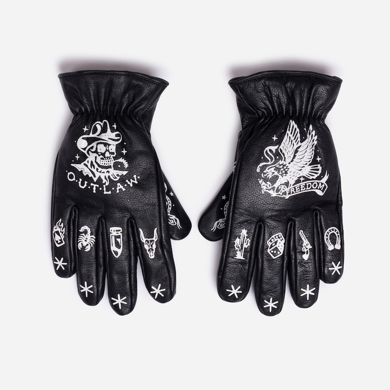 May include: Black leather gloves with white graphic designs. One glove features a skull with a cowboy hat and the word "OUTLAW". The other glove displays an eagle and the word "FREEDOM". Additional small graphics include a scorpion, cactus, and horseshoe.