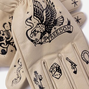 May include: Cream-colored leather gloves with black tattoo-style designs. The designs include an eagle with the word "FREEDOM", a skull, a snake, a cactus, dice, a revolver, a horseshoe, and stars.