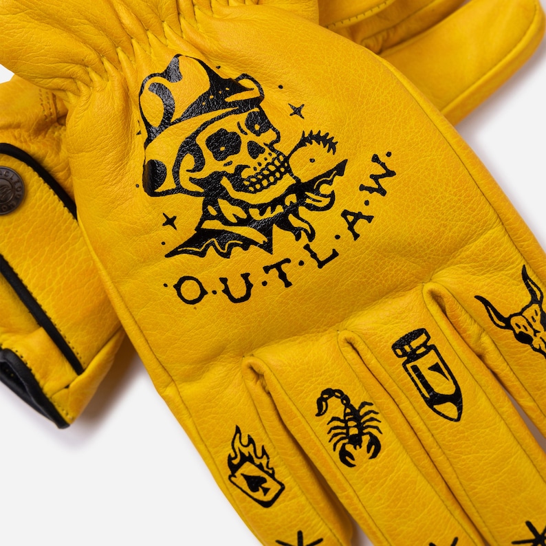 May include: Yellow leather work gloves with black graphic designs. The gloves feature a skull wearing a cowboy hat, the text "O.U.T.L.A.W.", a scorpion, a bomb, and other tattoo-style illustrations.