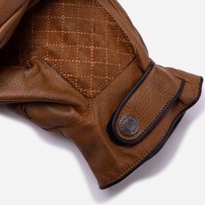 May include: Close-up of a brown leather glove with a quilted diamond pattern on the back of the hand. The glove has a black trim and a metal button on the wrist. The leather appears worn.