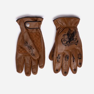 May include: Brown leather gloves with black stitched trim and a snap closure. The gloves feature black tattoo-style designs, including a snake, eagle with the word "FREEDOM", cactus, and other symbols. The gloves are designed for riding.
