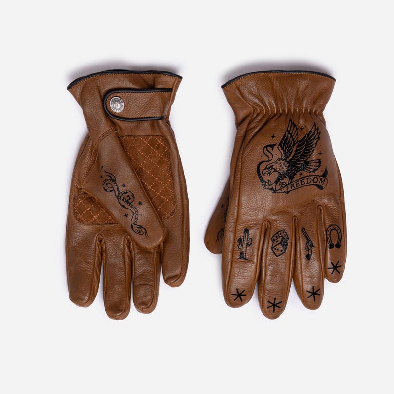 May include: Brown leather gloves with black stitched trim and a snap closure. The gloves feature black tattoo-style designs, including a snake, eagle with the word "FREEDOM", cactus, and other symbols. The gloves are designed for riding.