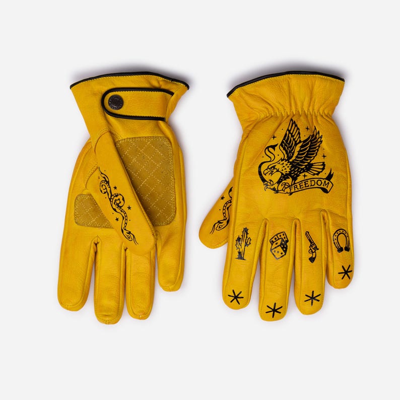 May include: A pair of yellow leather gloves with black trim and decorative stitching. The gloves feature black illustrations of a snake, eagle, cactus, dice, gun, horseshoe, and stars. The word "FREEDOM" is printed on one glove.