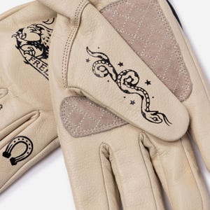 May include: Cream-colored leather gloves with black graphic designs. One glove features a snake and stars, the other a horseshoe and the word "FREE". The gloves have quilted beige patches on the palm and back of the hand.