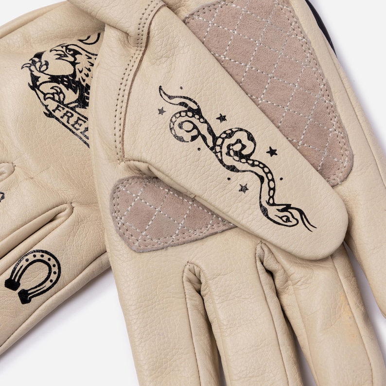 May include: Cream-colored leather gloves with black graphic designs. One glove features a snake and stars, the other a horseshoe and the word "FREE". The gloves have quilted beige patches on the palm and back of the hand.
