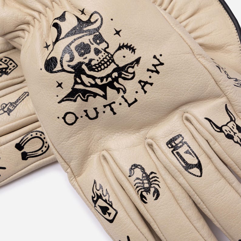 May include: Cream-colored leather gloves with black tattoo-style illustrations. The design includes a skull wearing a cowboy hat, the word "O.U.T.L.A.W.", a horseshoe, a scorpion, and other symbols. The gloves have a classic work glove design.