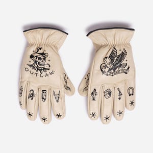 May include: A pair of cream-colored leather gloves with black graphic designs. One glove features a skull wearing a cowboy hat with the word "O.U.T.L.A.W." The other glove has an eagle with the word "FREEDOM". Additional small tattoos are on the fingers.