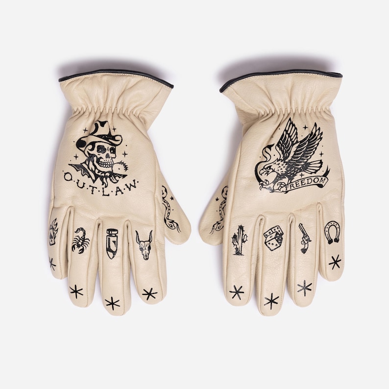 May include: A pair of cream-colored leather gloves with black graphic designs. One glove features a skull wearing a cowboy hat with the word "O.U.T.L.A.W." The other glove has an eagle with the word "FREEDOM". Additional small tattoos are on the fingers.