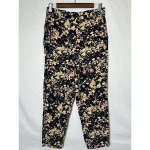 May include: Black cropped pants with a floral pattern in yellow and pink. The pants are hanging on a wooden hanger. The floral print is scattered across the fabric.
