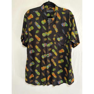 Könnte beinhalten: Schwarzes kurzärmeliges Hemd mit einem lebendigen Ananas- und Blättermuster. Das Hemd hat eine Brusttasche und einen klassischen Kragen. Das farbenfrohe Design umfasst gelbe, orangefarbene, grüne und blaue Ananas und Blätter.