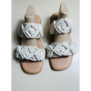 Puede incluir: Un par de sandalias blancas de crochet con suela plana. Las sandalias tienen dos bandas de material de crochet en la parte superior del pie.