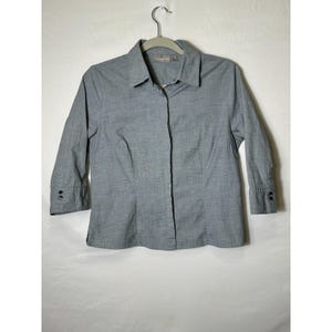 May include: A gray button-down shirt with three-quarter sleeves, black buttons, and a collar. The shirt is hanging on a hanger against a white background. The fabric has a subtle checkered pattern.