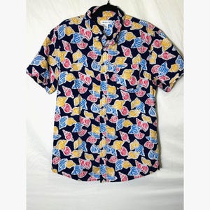 May include: Short-sleeved button-down shirt with a navy blue background and a colorful seashell pattern in yellow, pink, and blue. The shirt has a chest pocket and a collar. The brand name "RAIL" is visible on the collar.