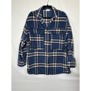 Puede incluir: Chaqueta de camisa a cuadros azul con cuello, cierre de botones y dos bolsillos en el pecho. La chaqueta presenta un patrón de cuadros clásico en tonos azules, marrones y blancos. La camisa está colgada de una percha negra.